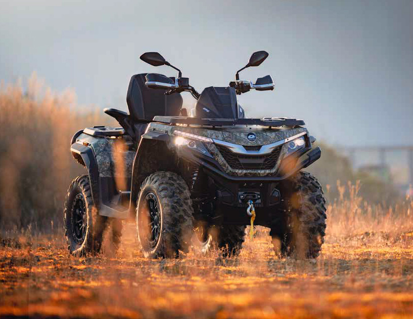 Camouflage-gepulltes QJ Motor Quad steht im Gras, im Hintergrund leuchten die Sonnenstrahlen eines tief stehenden Sonnenaufgangs oder -untergangs.