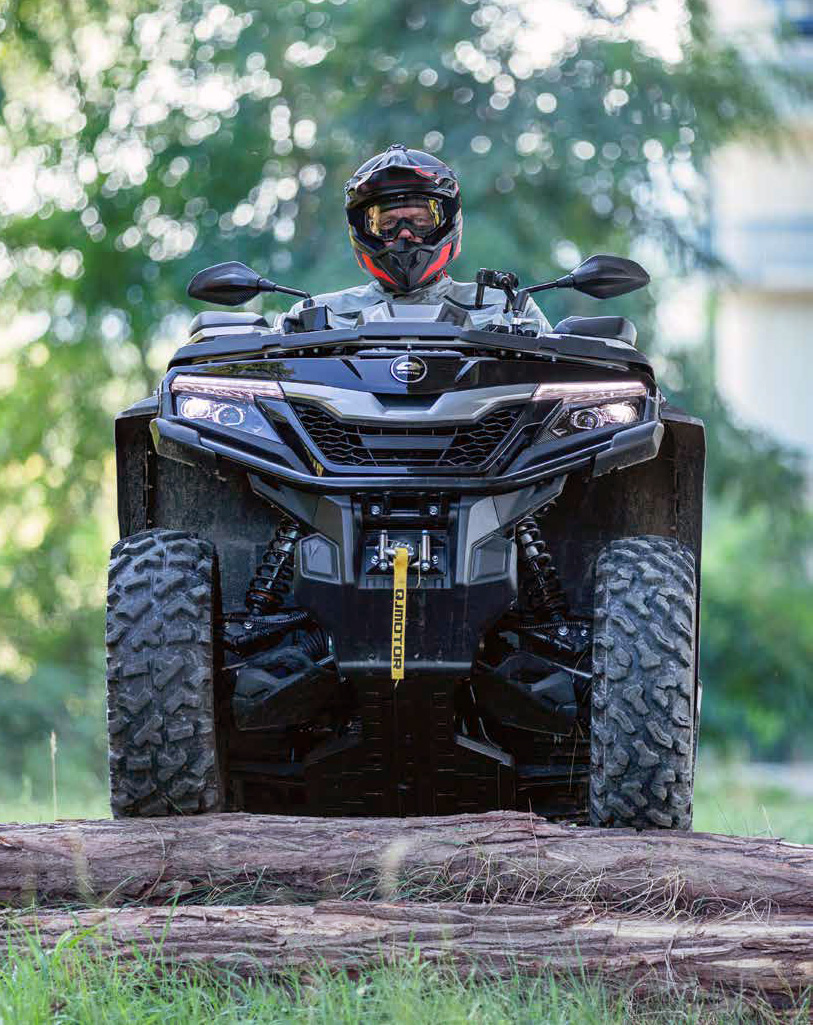 Frontalansicht eines QJ Motor Quads, das mit Fahrerhelm bekleidet über einen umgestürzten Baumstamm in einer grünen Waldumgebung fährt.