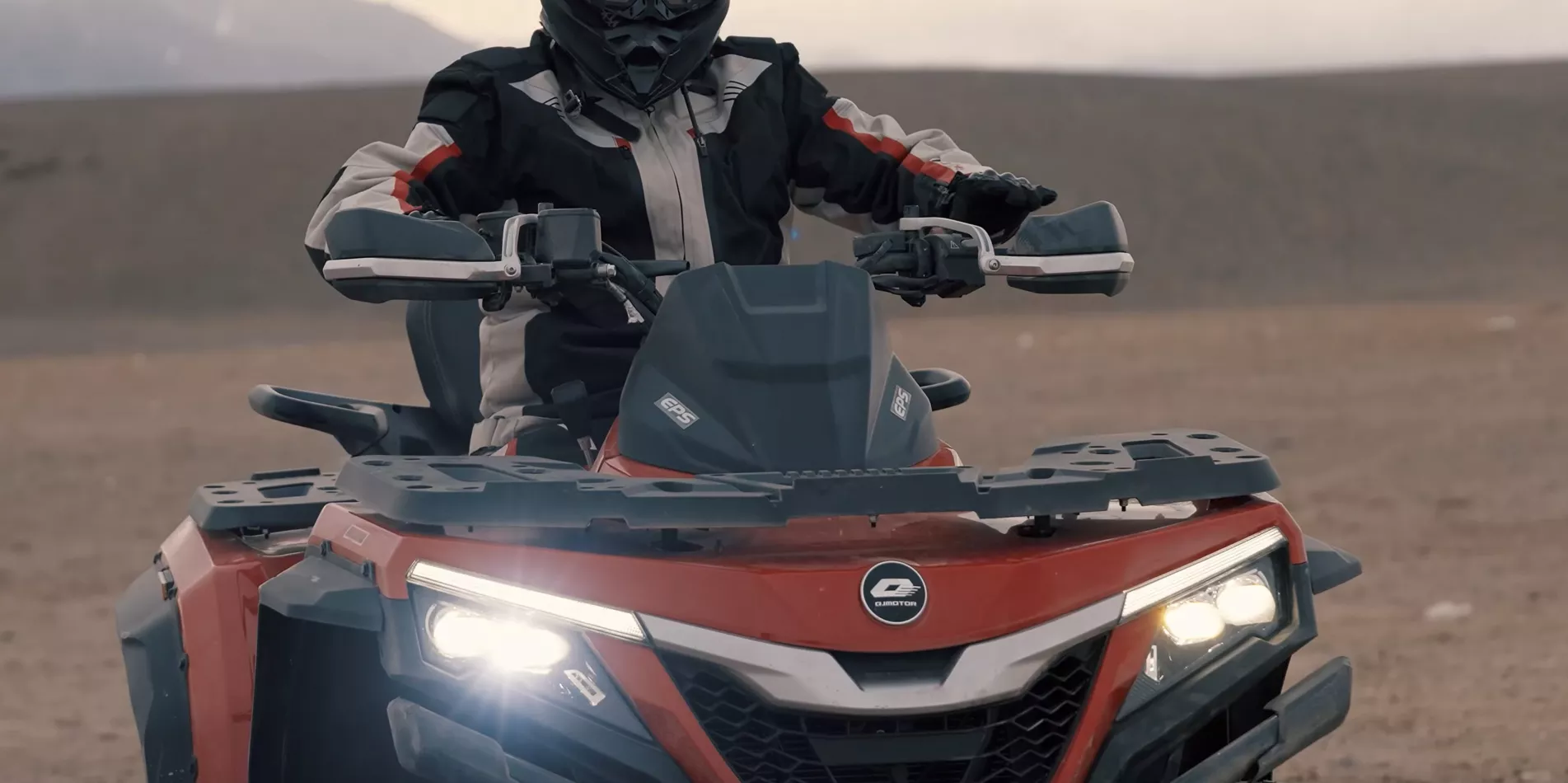 Ein rotes QJ Motor Quad steht bei Sonnenaufgang auf einer weiten, grasbewachsenen Ebene mit imposanten Bergen im Hintergrund.